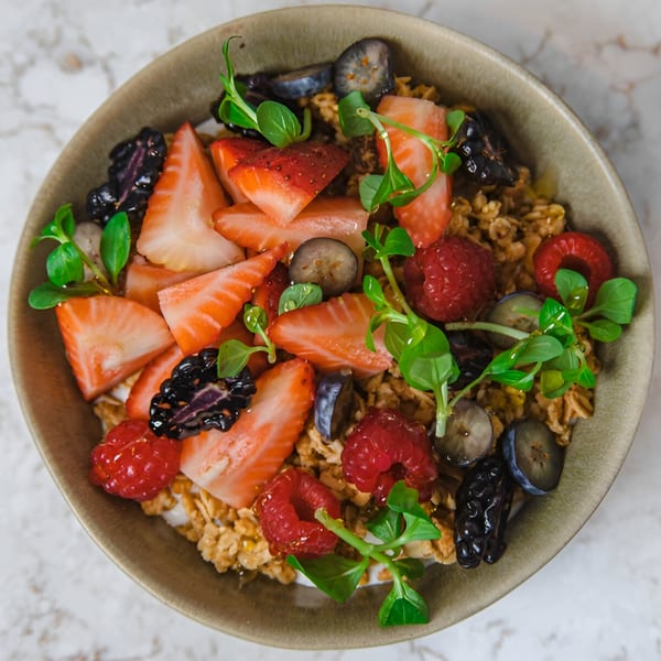 A bowl of granola with fresh strawberries, raspberries, blueberries, and microgreens on a marble surface.