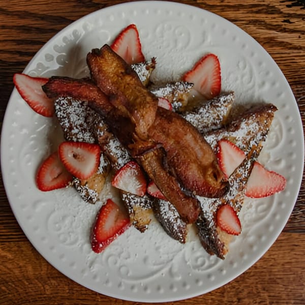 French toast with powdered sugar, sliced strawberries, and bacon on a white plate.