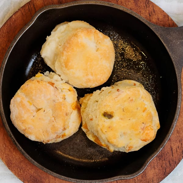Three golden-brown biscuits, one with visible cheese, served in a black cast-iron skillet on a wooden board.